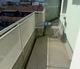 Balcony with tiled floor, metal railing, small round table, and folding chair, overlooking cityscape with rooftops and mountains.