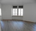 Empty room with wooden flooring, white walls, and large windows with a view of a building outside.