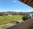 Balcony with wooden railings and a view of a rural landscape, including a green field with grazing cows, scattered houses, and a clear blue sky.