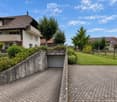 Two-story house with a tiled roof, white exterior, and a balcony on the upper floor. The house features a driveway, a garage entrance, and is surrounded by greenery and a well-maintained lawn.