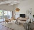 Modern living room with large windows and balcony access, featuring a grey sofa, coffee table, wooden chairs, TV, floor lamp, bookshelf, and a plant.