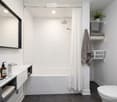 Modern bathroom with white tiled walls, dark grey floor, white bathtub, shower curtain, and a sink with a mirror above it. Shelves hold towels, a laundry basket, and a small plant. A toothbrush and soap dispenser are on the sink counter.
