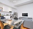 Open-plan living and dining area with a grey sofa, glass dining table, and modern kitchen featuring stainless steel appliances, white cabinets, and a countertop with various items.