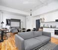 Modern, open-concept studio apartment with a grey sofa, dining table, bed, and a compact kitchen area featuring white cabinets and stainless steel appliances.
