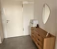 A small, neatly furnished room with a wooden dresser, a round mirror, and a small cabinet. The room has a tiled floor and white walls.