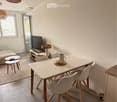 Bright, modern dining and living area with white walls, tiled floor, and large window. Features a white dining table with four chairs, a sofa, coffee table, and a television on a wooden stand.
