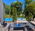 Well-maintained garden with a swimming pool, patio furniture including a sofa, coffee table, and chairs, surrounded by lush greenery and trees.