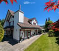 Two-story house with red-tiled roof, white and red facade, chimney, and well-maintained garden with lawn, patio, and outdoor seating area.