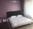 Modern bedroom with a large bed, white bedding, nightstand, and a grey rug on the floor. The walls are painted in a soft purple color, and there are dark curtains on the windows.