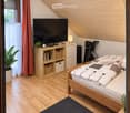 Cozy bedroom with wooden floors, slanted wooden ceiling, bed with patterned quilt, nightstand with lamp, bookshelf with books and baskets, potted plant, television, and a tripod in the corner.
