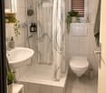 Modern bathroom with white tiles, a bathtub with a shower curtain, a sink with a mirror, a toilet, and decorative plants on the windowsill and shelf.