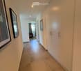 A clean, well-lit hallway with tiled flooring, white walls, built-in wardrobes, and framed artwork. A glimpse of a room with a chair and window is visible at the end.