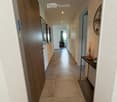 Clean, well-lit hallway with tiled flooring, white walls, and recessed lighting. Features a small console table with a vase and decorative items, framed artwork on the walls, and a glimpse of a living area at the end.