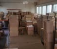 Large, empty warehouse space with numerous cardboard boxes stacked throughout, large windows with blinds, and a forklift in the center.