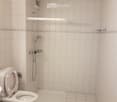 Small bathroom with white tiled walls and floor, featuring a toilet, a shower area with a handheld showerhead, and a small shelf above the shower.
