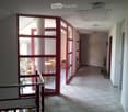 A well-lit hallway with tiled flooring, white walls, and red-framed glass doors leading to a room with wooden chairs and a table. A fire extinguisher is mounted on the wall.