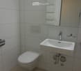 Modern bathroom with white tiled walls, a toilet, a sink with a modern faucet, a mirror with a built-in radiator, and a light switch on the wall.