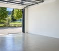 Empty garage with a large, open garage door leading to a grassy area with trees and a picnic table outside.