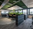 Modern office space with desks, chairs, computers, and large windows offering a scenic view. Features include grey carpeting, grey cabinets, and indoor plants.