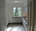 Empty kitchen with white walls, grey cabinets, a window with a view of greenery, and a radiator below the window.