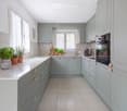 Modern kitchen with light blue cabinets, white countertops, and stainless steel appliances. Features a central island, built-in oven, and various kitchen items including potted plants and a coffee maker.