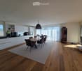 Open-plan kitchen and dining area with modern white cabinets, a wooden dining table with chairs, a wine rack, a sofa, and a coffee table. Large windows with curtains allow natural light to fill the space.