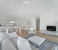 Modern, open-concept living and dining area with light grey furniture, white coffee table, grey sofa, armchair, dining table with chairs, and a television on a stand. The space features light wood flooring, white walls, and contemporary decor including po