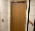 A narrow hallway with a wooden door and a staircase railing visible. The walls are concrete, and the floor is tiled.