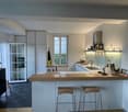 Modern kitchen with white cabinets, wooden bar counter, two stools, sink, oven, and various kitchen items on the counter and shelves.