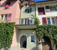 A charming multi-story house with pastel-colored facades, wooden shutters, and a balcony adorned with greenery and flowers. The entrance features a stone archway and a barrel planter.