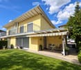 A modern two-story house with a yellow facade, featuring a covered patio area with a table and chairs, surrounded by a well-maintained lawn and garden.