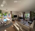 Bright living room with a white sofa, coffee table, dining area with chairs and table, fireplace, and large windows with plants.