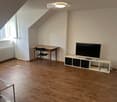Bright, slanted-ceiling room with wooden flooring, a TV on a white stand, a small wooden desk with a chair, and a glass coffee table.