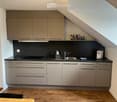 Modern kitchen with light grey cabinets, black countertops, stainless steel sink, and various appliances. Wooden floor and a small dining area visible.