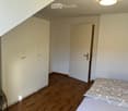 A small, furnished room with a wooden floor, a bed with a patterned bedspread, a white wardrobe, and a wicker chair.