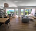 Bright living and dining area with wooden flooring, large windows offering a scenic view, a grey sofa, a coffee table, a dining table with chairs, a shelf with plants and decor, and a television on a stand.