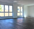 Empty room with light-colored walls and wooden flooring, featuring large windows with views of rooftops and trees.