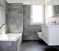 Modern bathroom with grey tiled walls, a white bathtub, a white sink with a chrome faucet, a toilet, and a mirrored cabinet.