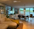 Open-plan living and dining area with a sofa, coffee table, dining table, chairs, pendant lights, and a large window overlooking a garden.