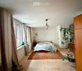 Cozy bedroom with wooden floor, large window with curtains, bed with patterned bedding, nightstand, and plants.