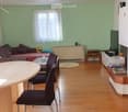 A cozy living room with a sofa, a dining table with chairs, a television, a small side table, and a plant. The room features light green walls, wooden flooring, and large windows with white curtains.