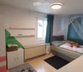A small, well-lit bedroom with a bed, wardrobe, shelves, and a table. The room features a window with curtains, a radiator, and a colorful accent wall.