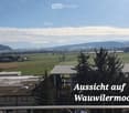 Panoramic view of a rural landscape with fields, scattered houses, and distant mountains under a clear sky.