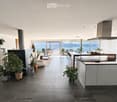 Open-plan living and kitchen area with large windows offering a scenic view, featuring a sofa, coffee table, kitchen island, bookshelf, and potted plants.