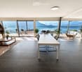 Modern open-plan living and dining area with large windows offering a scenic mountain view, featuring a white dining table, grey chairs, a white sofa, a wooden coffee table, and several potted plants.