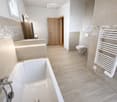 Modern bathroom with a white bathtub, toilet, radiator, and wooden door. Light-colored tiles and wooden flooring.