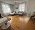 Modern living room with wooden flooring, large windows, grey sectional sofa, coffee table, cat tree, and decorative items.