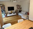 Modern living room with light wooden flooring, a white sofa, a wooden dining table with two chairs, a TV mounted on the wall, and several bookshelves filled with books and decorative items.