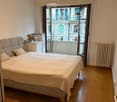 Bedroom with wooden flooring, large bed with pillows and blanket, nightstand with lamp, radiator, and open sliding doors leading to a balcony.