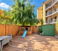 Wooden deck with a table, chairs, slide, small stool, and a basketball hoop, surrounded by a wooden fence and trees.